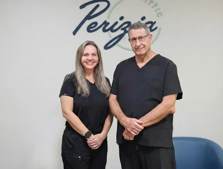 Two staff members in black scrubs posing before the Perizia Aesthetics sign.