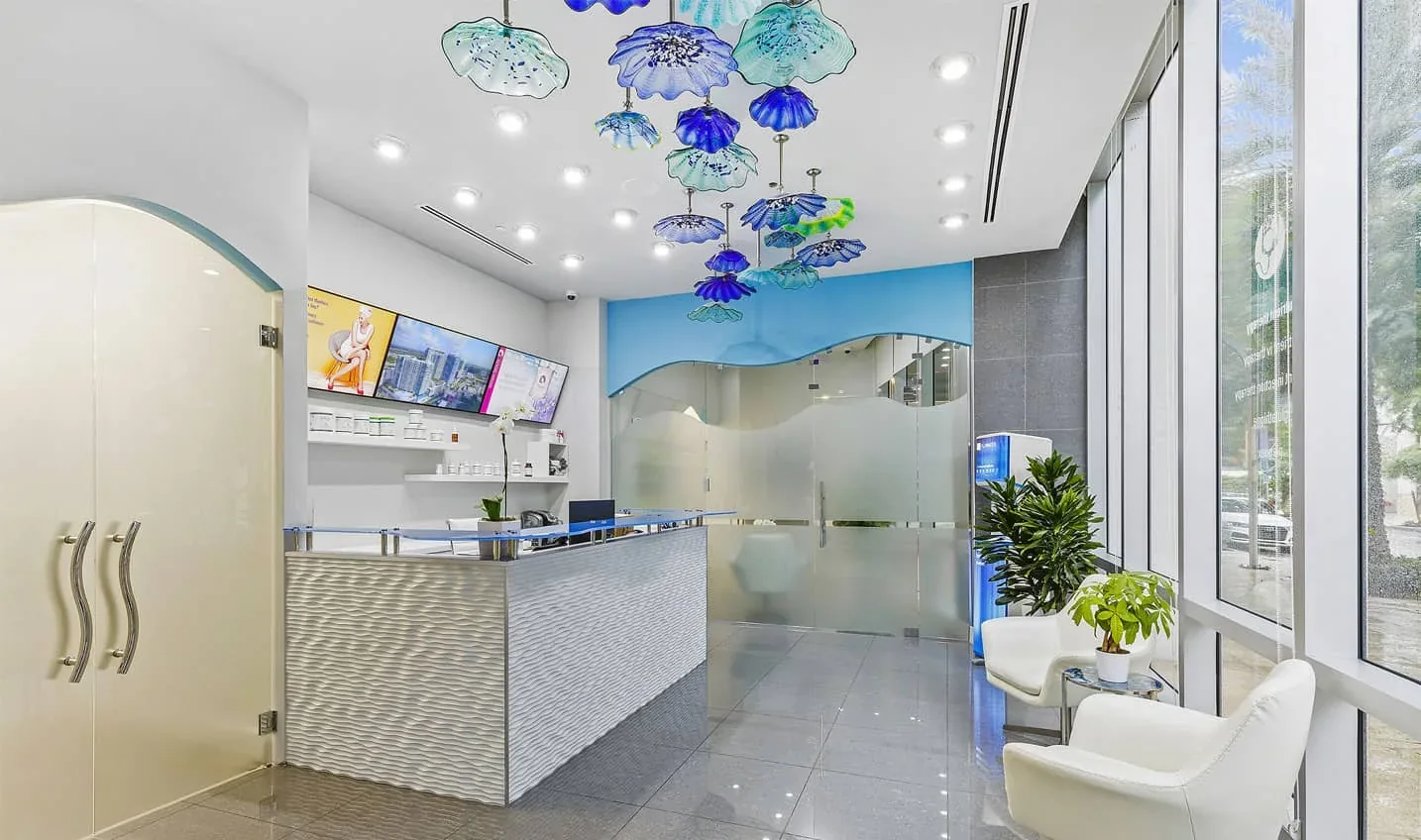 Empty reception desk and white waiting chairs beneath blue glass ceiling fixtures.