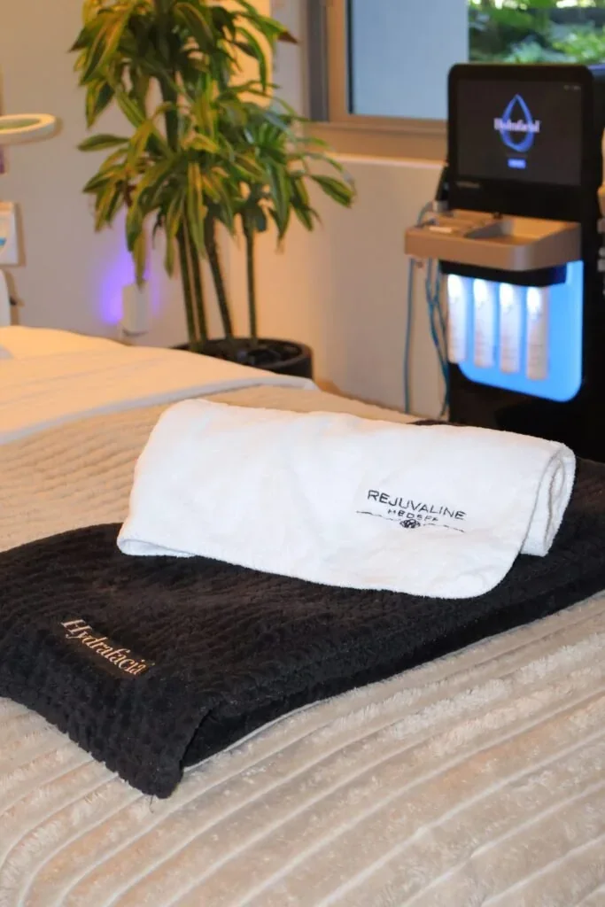 Folded Rejuvaline towels beside a Hydrafacial unit and a plant.
