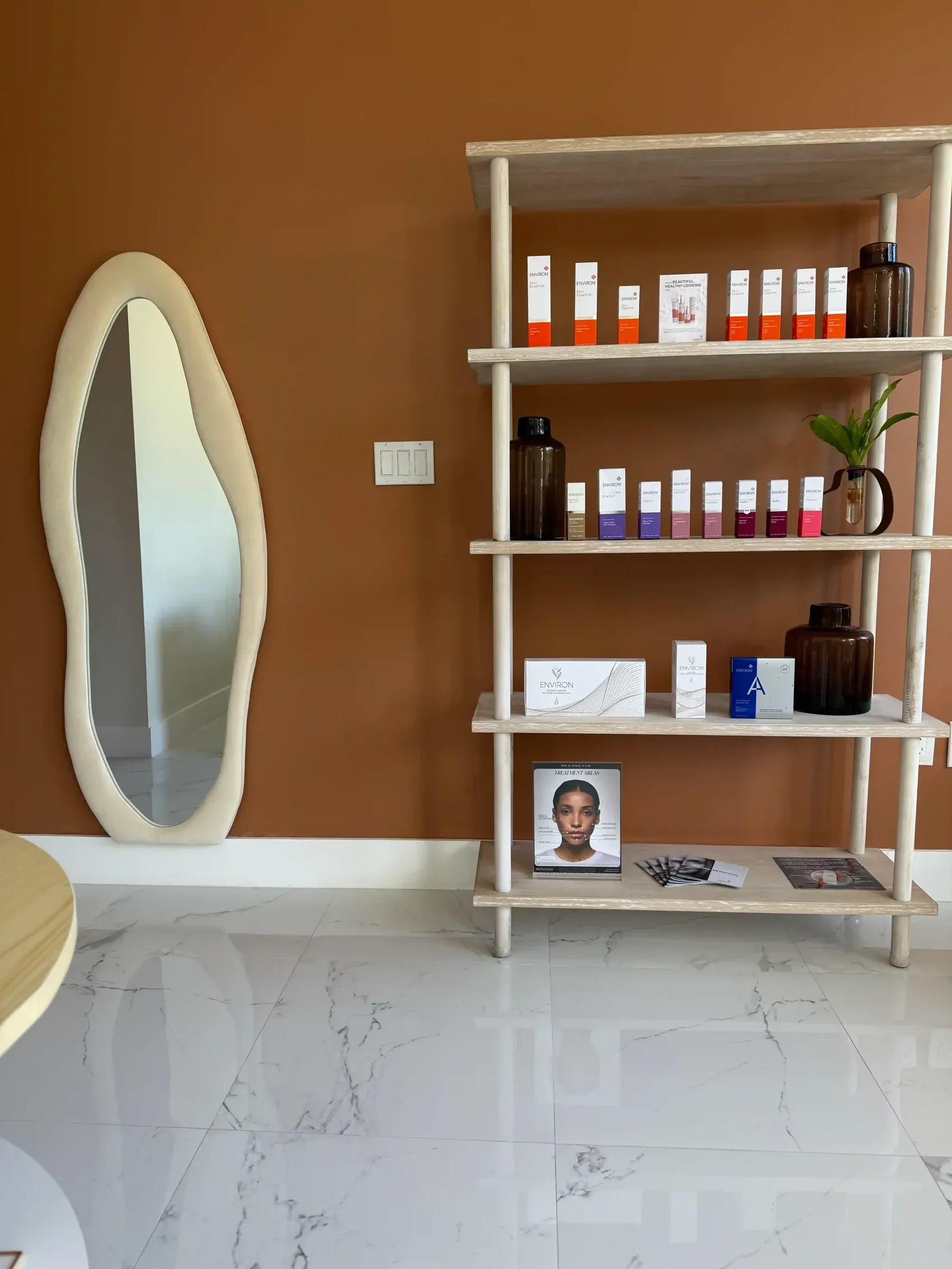 Skincare shelves and a tall oval mirror against a rust-colored wall.