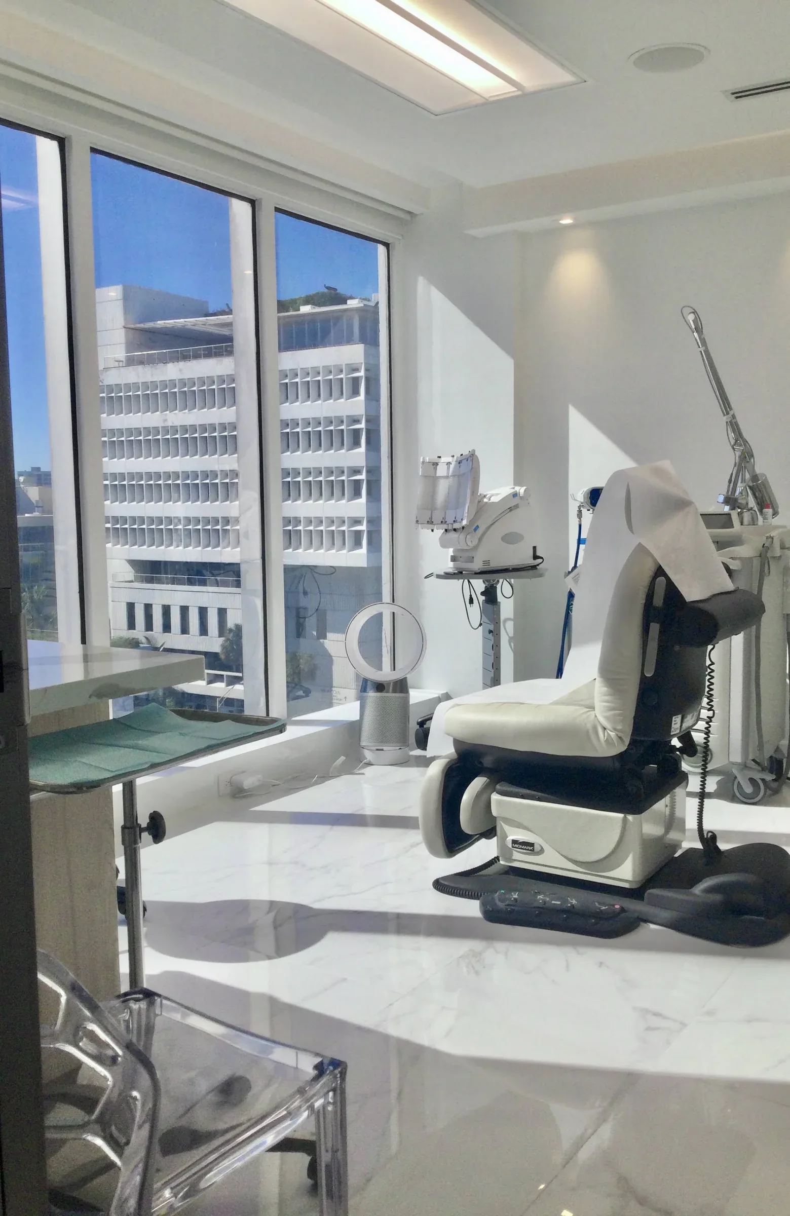 Empty examination chair beside tall windows overlooking nearby Miami Beach buildings.