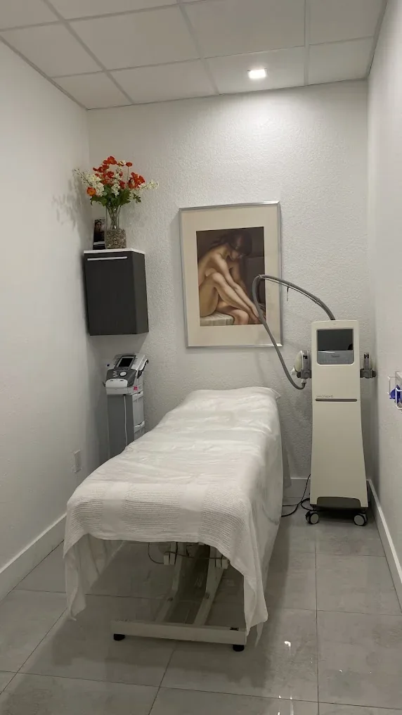 Narrow empty spa room with white covered bed, equipment and framed wall art.