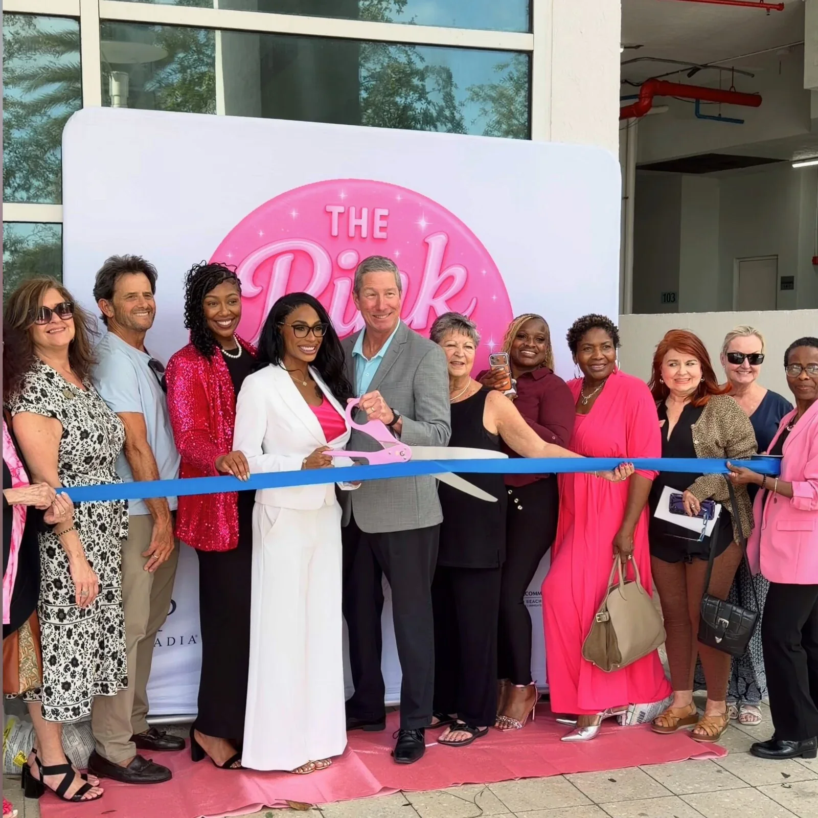 A group gathered for a ribbon-cutting in front of The Pink Spot's branded backdrop.