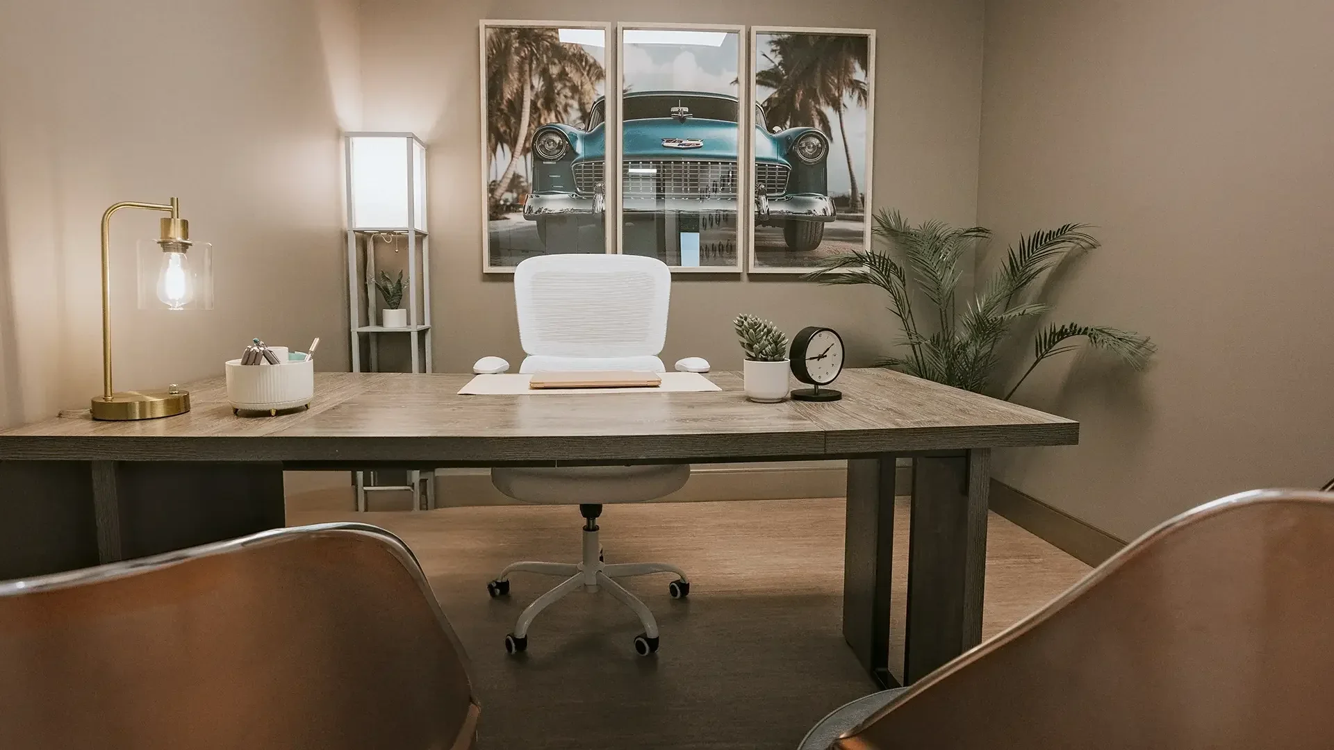 Desk, white chair, plants and framed blue-car artwork in an empty office.