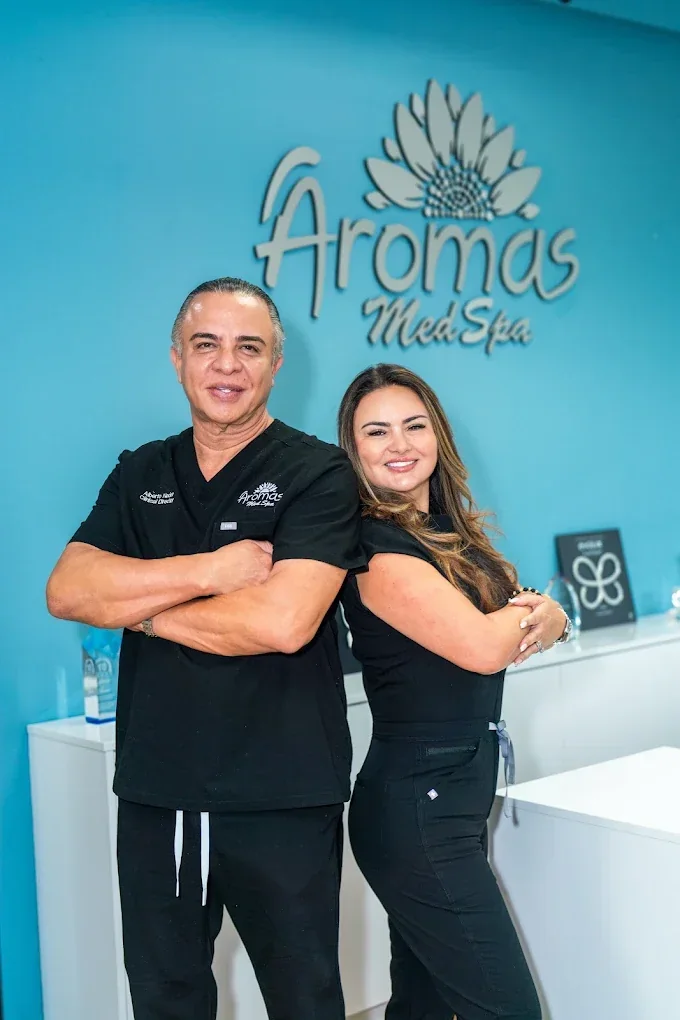 Two staff members in black uniforms posed in front of a turquoise Aromas Med Spa sign.