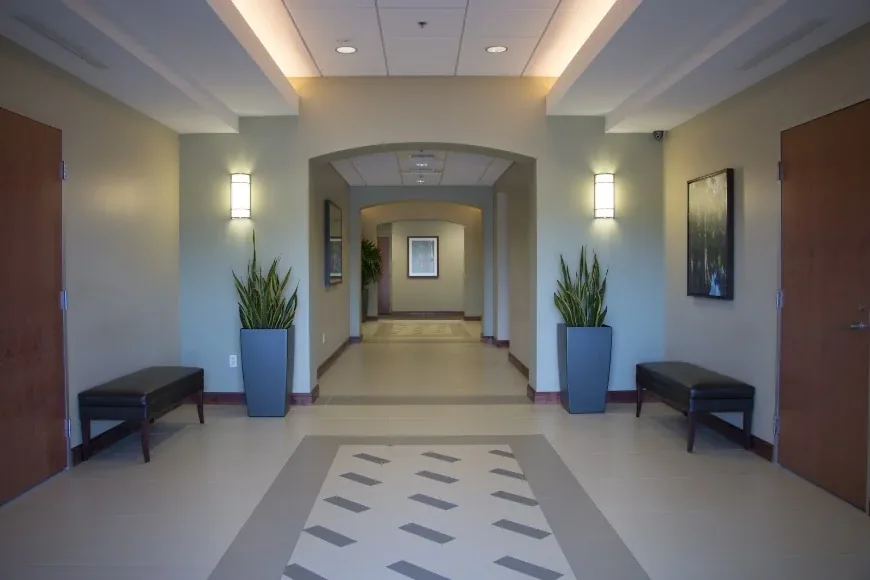 Empty medical-office corridor with tiled floor, benches, wall lights and tall potted plants.